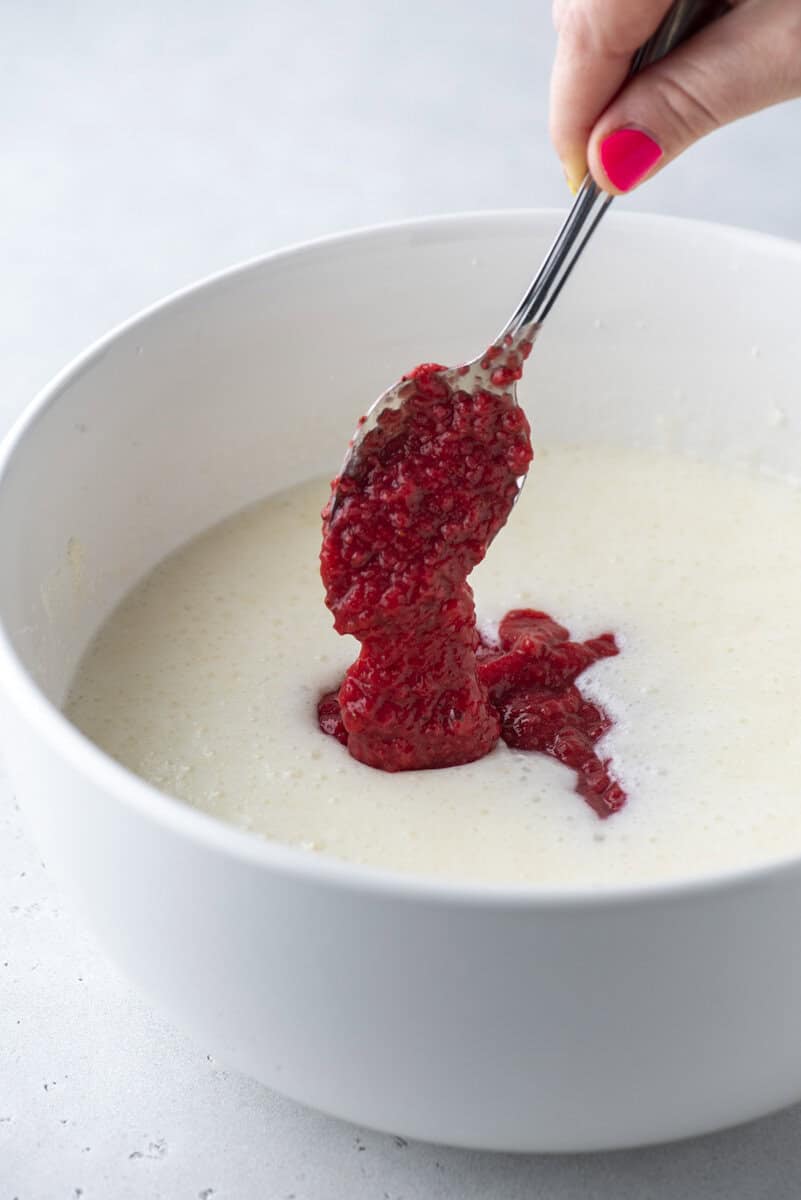 A hand holding a spoon with bright pink nail polish adds mashed raspberries to a bowl of pale batter, preparing to mix the ingredients together.