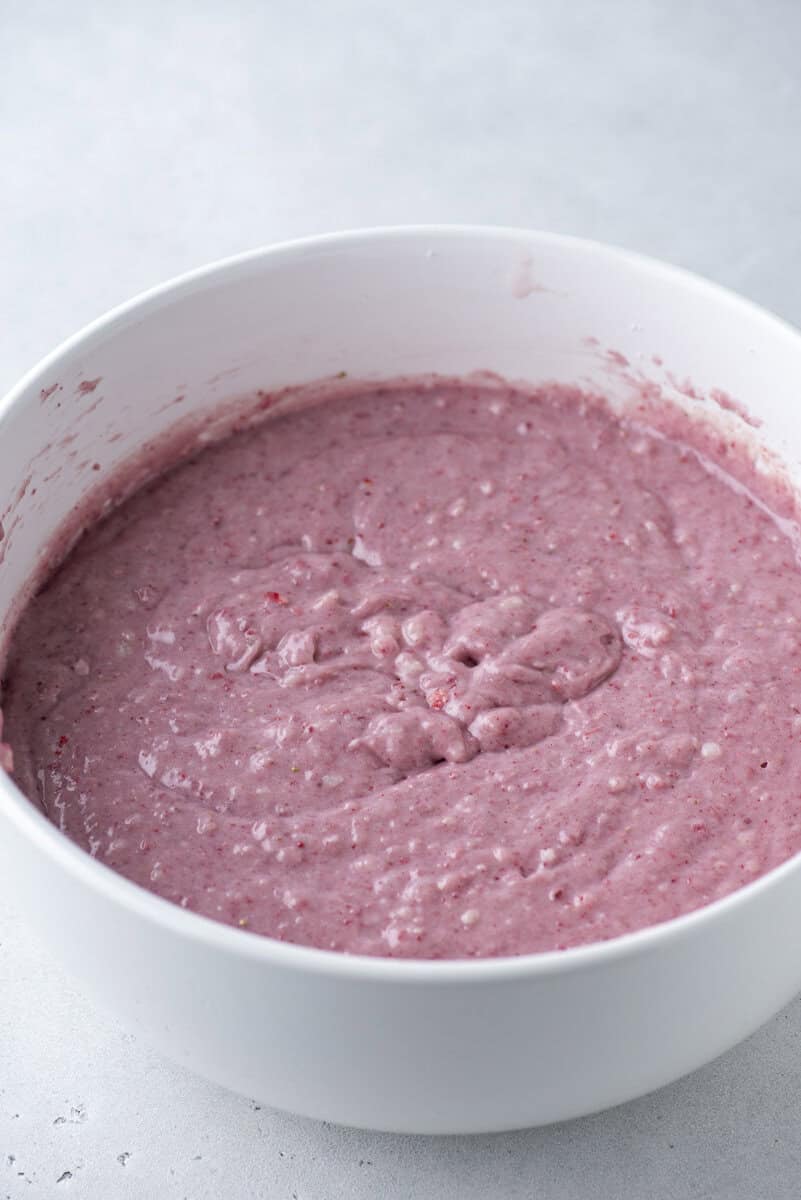 A white bowl filled with thick, pink batter, likely mixed with berries or fruit, sits on a light gray surface.