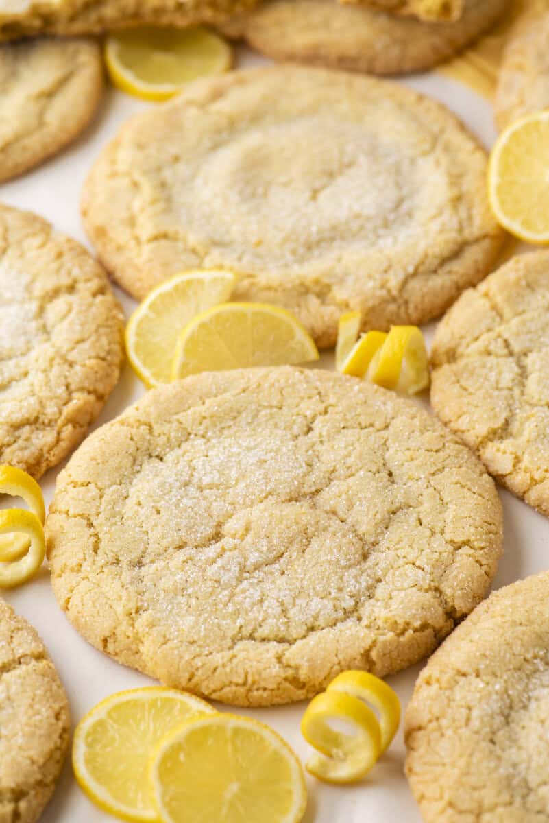 Large, round lemon sugar cookies are arranged on a surface with a light dusting of sugar on top. Lemon slices and lemon twists are scattered around, adding a fresh, bright accent.