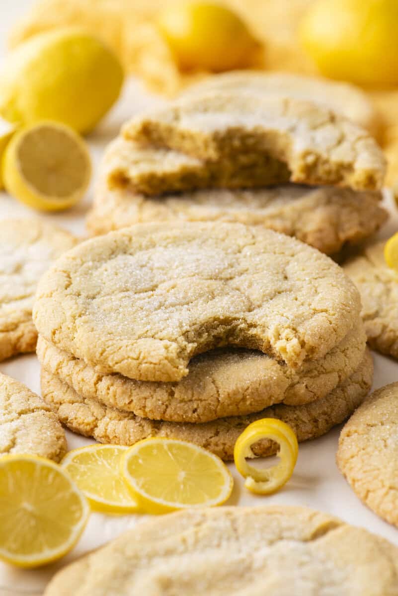 A stack of sugar-coated lemon cookies, with one cookie on top showing a bite taken out. Fresh lemon slices and whole lemons are scattered around the cookies on a light surface.
