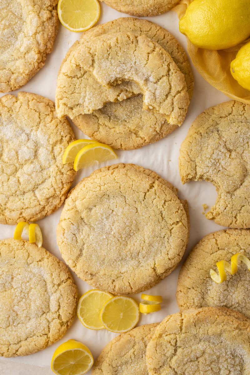 Large, round lemon sugar cookies are arranged on a light surface with lemon slices and whole lemons scattered around. One cookie has a bite taken out of it. The cookies have a crinkled texture and a sugar-coated top.