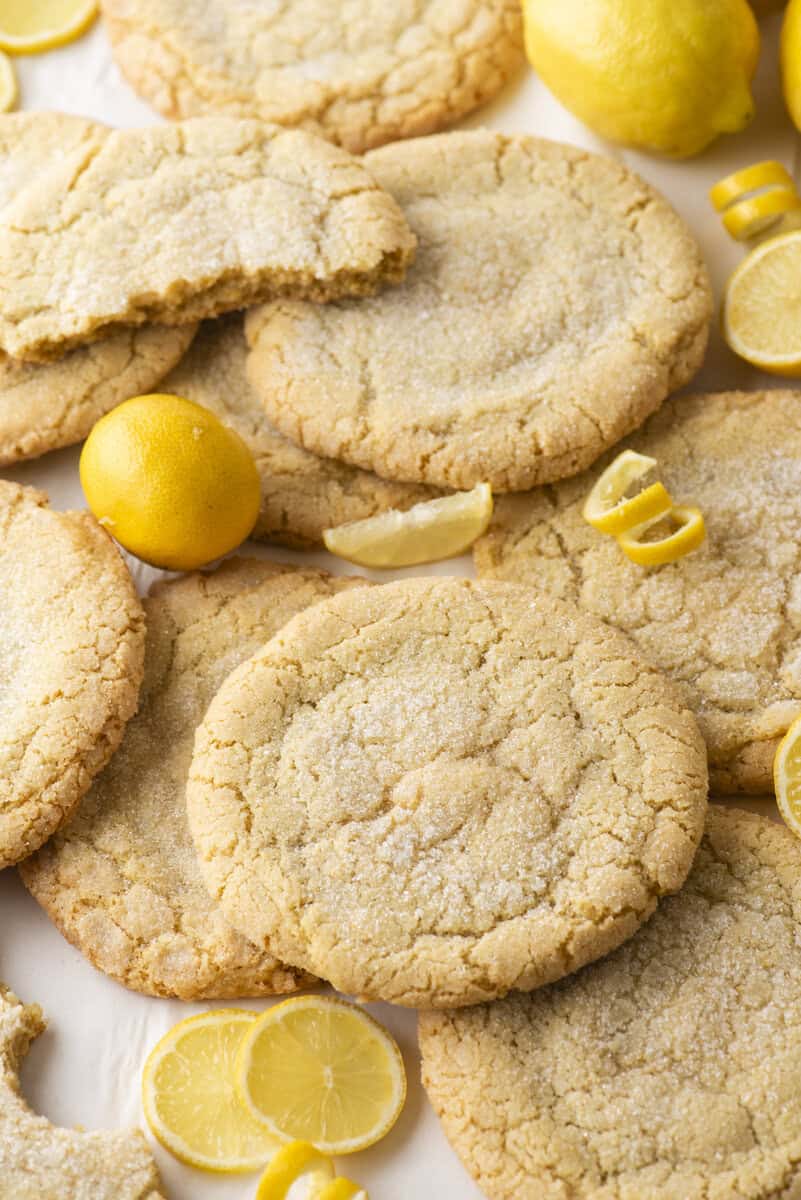 Large, round lemon sugar cookies are arranged on a surface with fresh whole and sliced lemons scattered around. One cookie is partially eaten, showing the soft, crumbly interior.