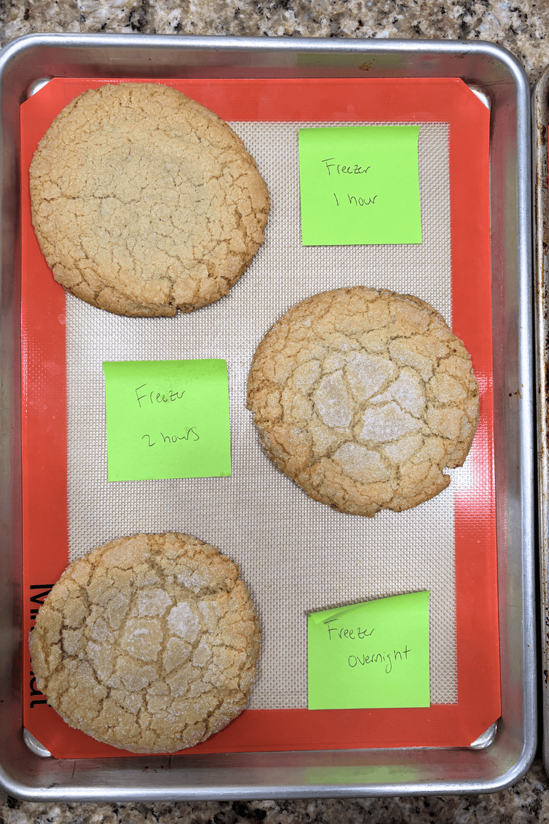 A baking tray with four large cookies on a silicone mat, each labeled with a green sticky note: Freezer 1 hour, Freezer 2 hours, Freezer 1 hour, and Freezer Overnight.