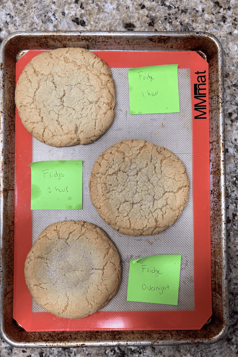 A baking tray with four large cookies on a silicone mat. Each cookie is labeled with a green sticky note: Fridge 2 hours, Fridge 1 hour, Fridge Overnight, and Fridge. The tray is on a granite countertop.