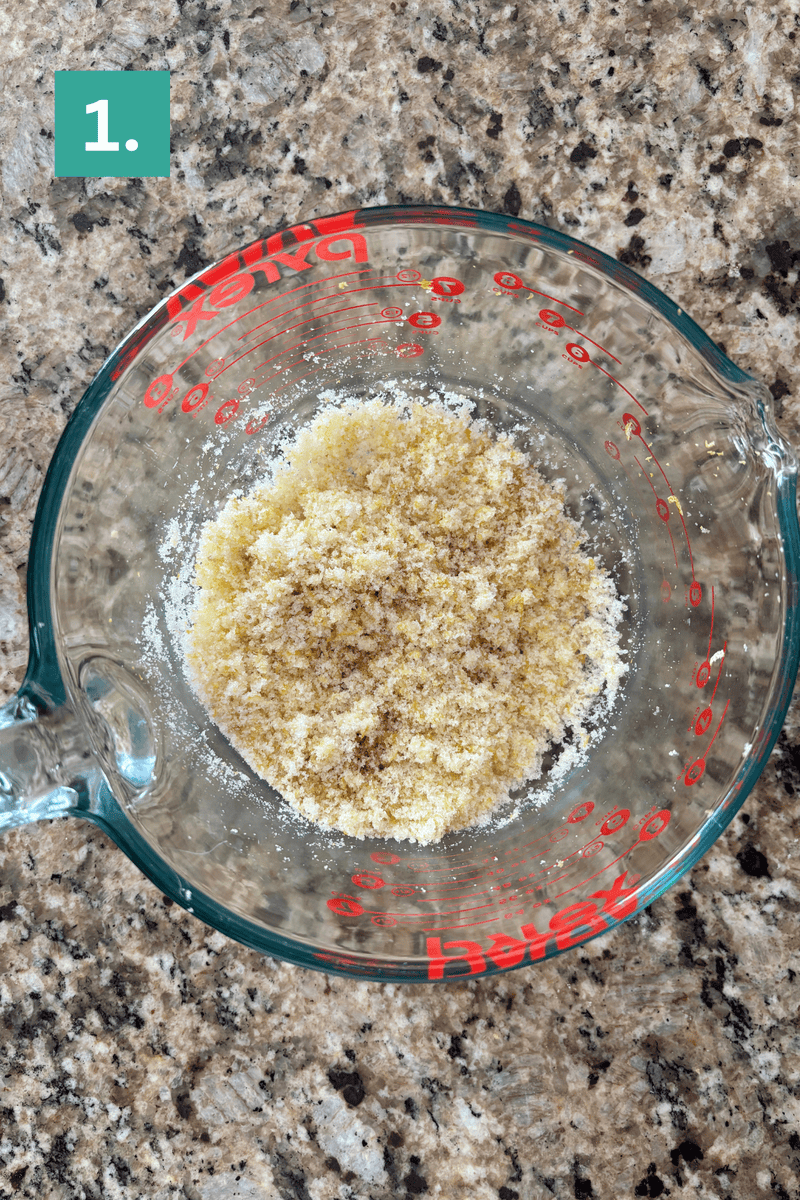 A glass measuring cup with a mixture of dry, crumbly ingredients sits on a speckled brown countertop. A green box with the number 1 is in the top left corner, indicating the first step.