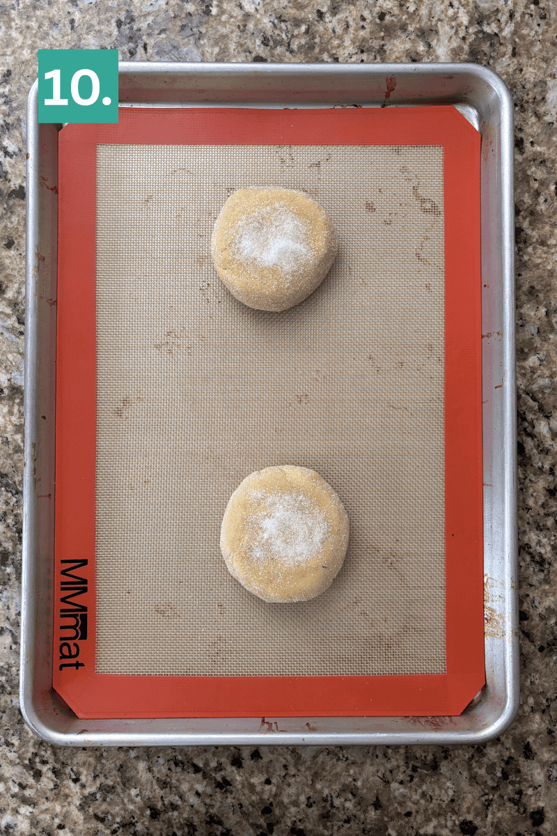 A baking sheet with a silicone mat holds two round, unbaked dough balls sprinkled with sugar. A green box with “10.” is in the top left corner. The tray is on a speckled countertop.