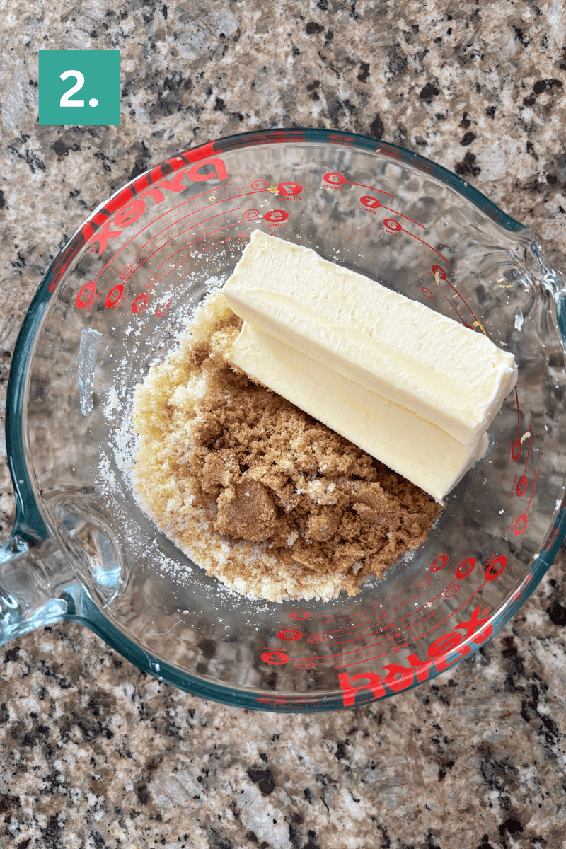 A glass measuring cup on a granite countertop contains two sticks of butter, brown sugar, and granulated sugar. A green box with the number 2 appears in the upper left corner.