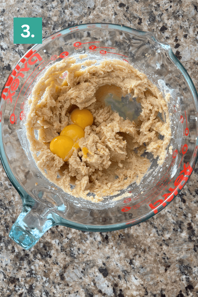 A glass measuring cup containing a creamy batter and three cracked eggs, sitting on a granite countertop. A green box with the number 3 is in the top left corner.