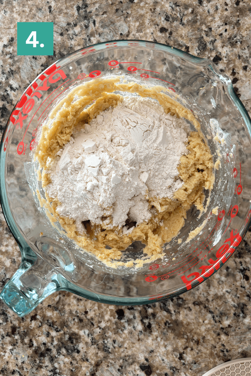 A glass measuring cup with cookie dough and flour being mixed, placed on a granite countertop. A green box with the number 4 is in the top left corner.