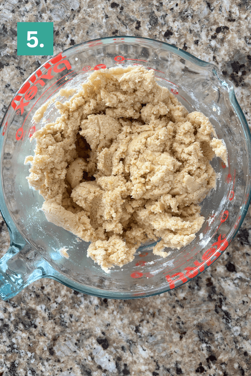 A glass measuring bowl filled with cookie dough sits on a speckled granite countertop. A green square in the top left corner displays the number 5 in white.
