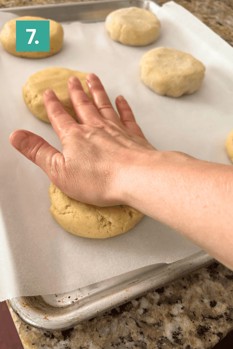 A hand presses down on a round ball of dough, flattening it on a parchment-lined baking sheet. Other unflattened dough balls are nearby. A green box with the number 7 is in the upper left corner.