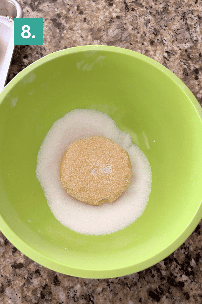 A round dough ball coated in sugar sits in the center of a green bowl on a granite countertop. A small label with the number 8 is in the upper left corner of the image.