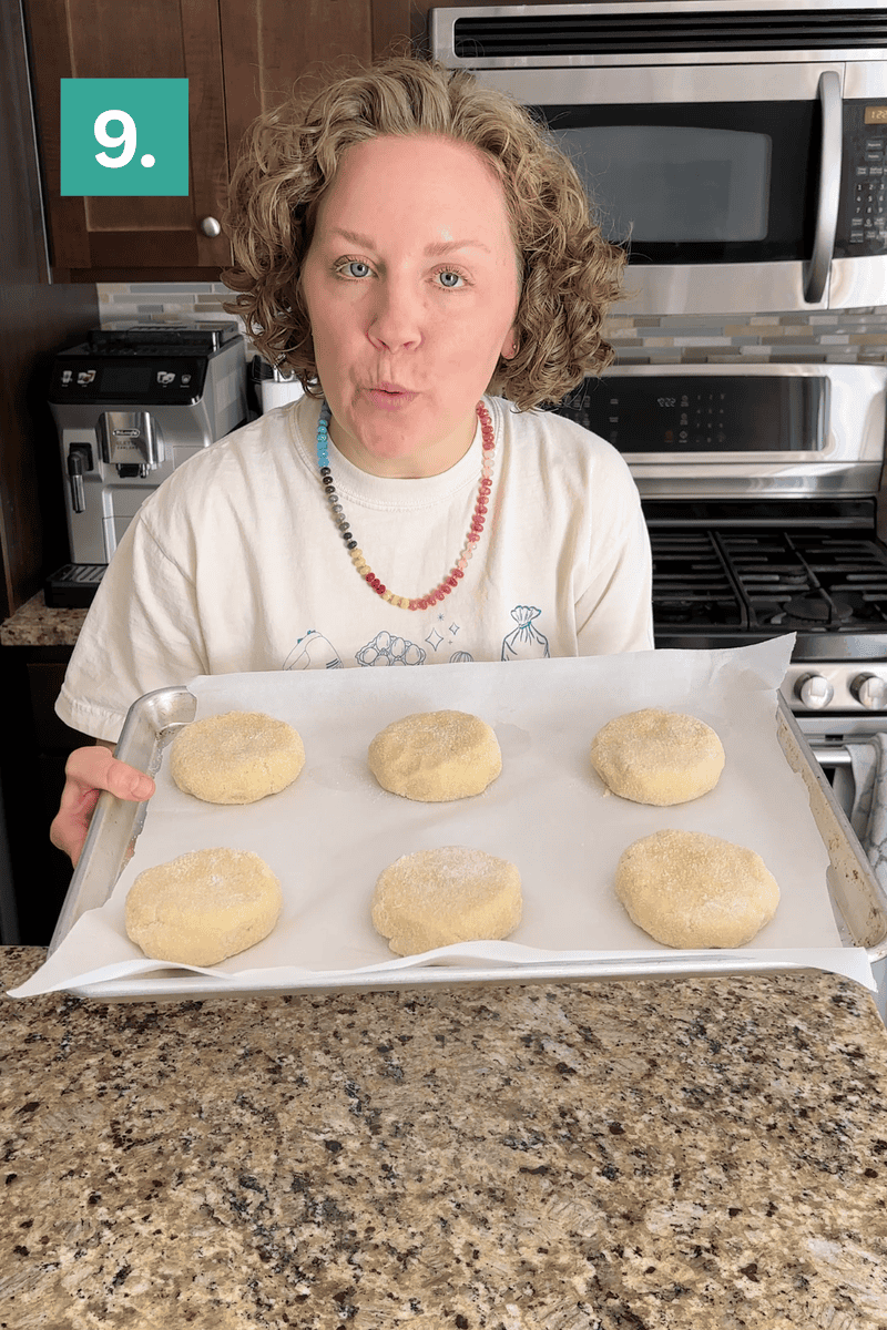 A person with curly hair holds a baking sheet with six sugar-coated dough rounds on parchment paper in a kitchen. A green box with the number 9 is in the top left corner of the image.