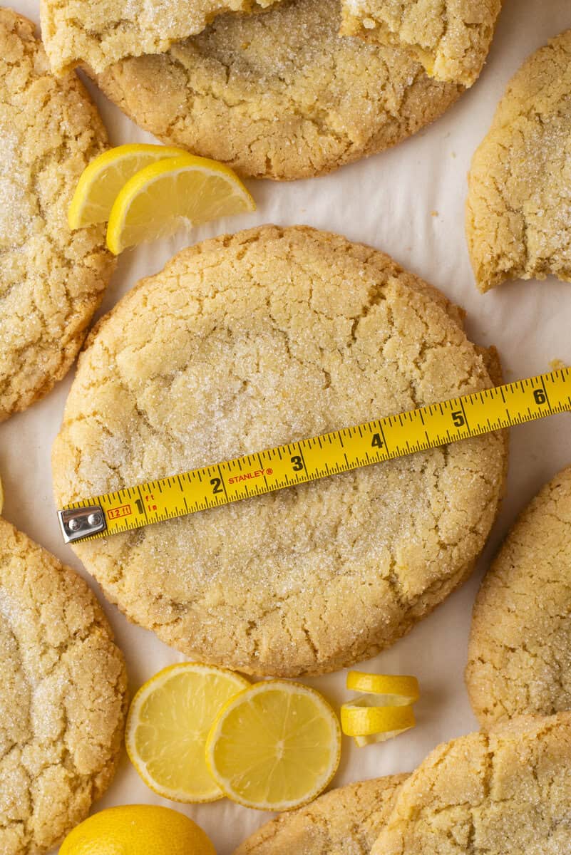 A large lemon cookie sprinkled with sugar is measured with a yellow tape measure. The cookie is surrounded by sliced lemons and other similar cookies on a parchment-lined surface.