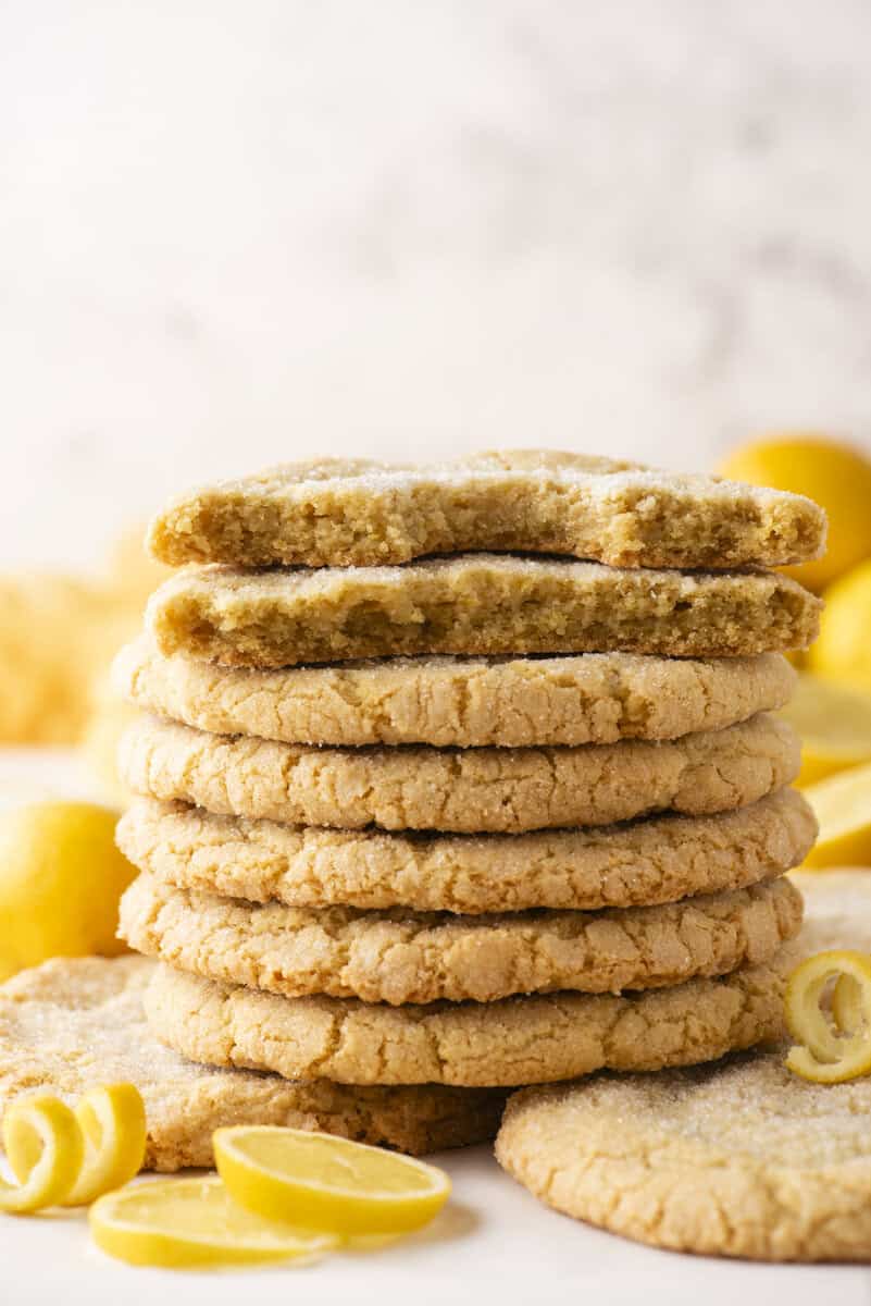 A stack of sugar-coated lemon cookies is shown, with the top cookie broken in half to reveal a soft, crumbly interior. Lemon slices and lemon zest curls are scattered around the cookies.