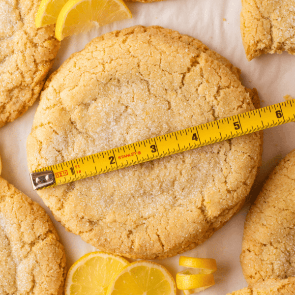 A large, round lemon sugar cookie topped with sugar is measured by a yellow tape measure. Lemon slices and other cookies are visible around it on a light surface.