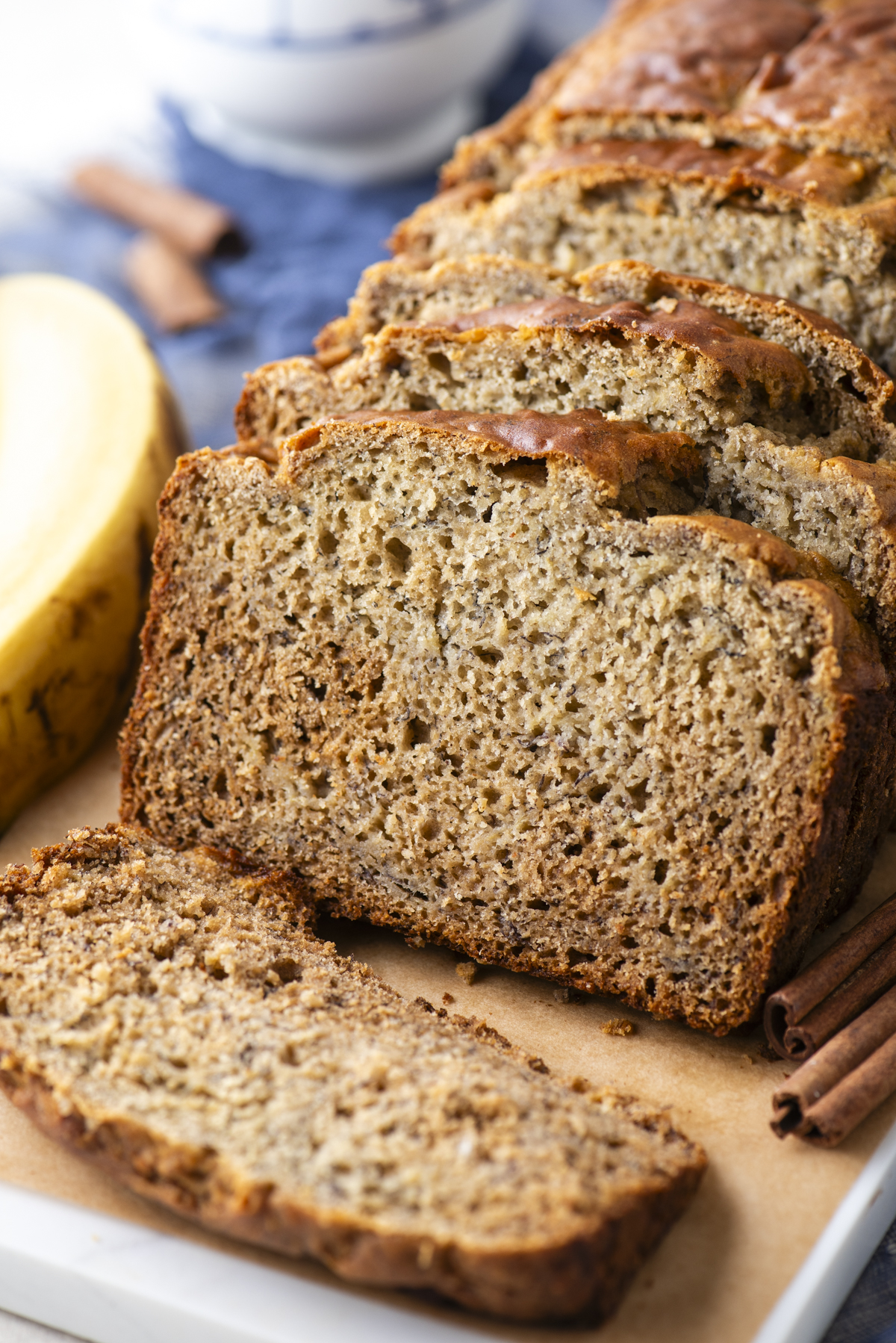 A close-up of sliced banana bread on a cutting board with a banana and cinnamon sticks nearby. The bread looks moist and has a golden-brown crust.