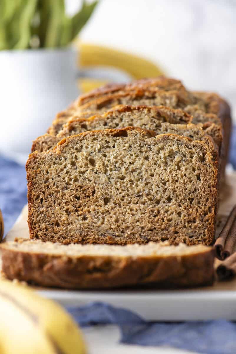 Close-up of several slices of banana bread arranged in a row on a white plate, with blurred bananas and a green plant in the background.