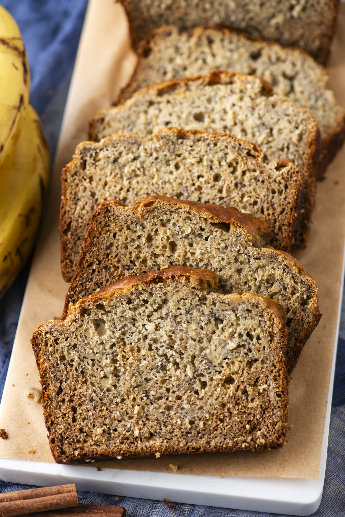 Sliced banana bread on a rectangular white plate, with a bunch of bananas beside it. The bread has a golden-brown crust and a moist, speckled interior. The scene is set on a blue cloth.
