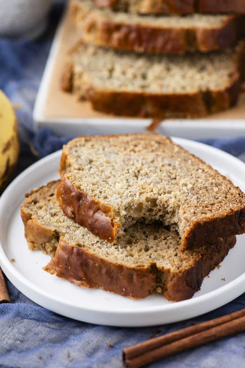 Two slices of banana bread on a white plate, one with a bite taken out, with more slices in the background and a cinnamon stick nearby.