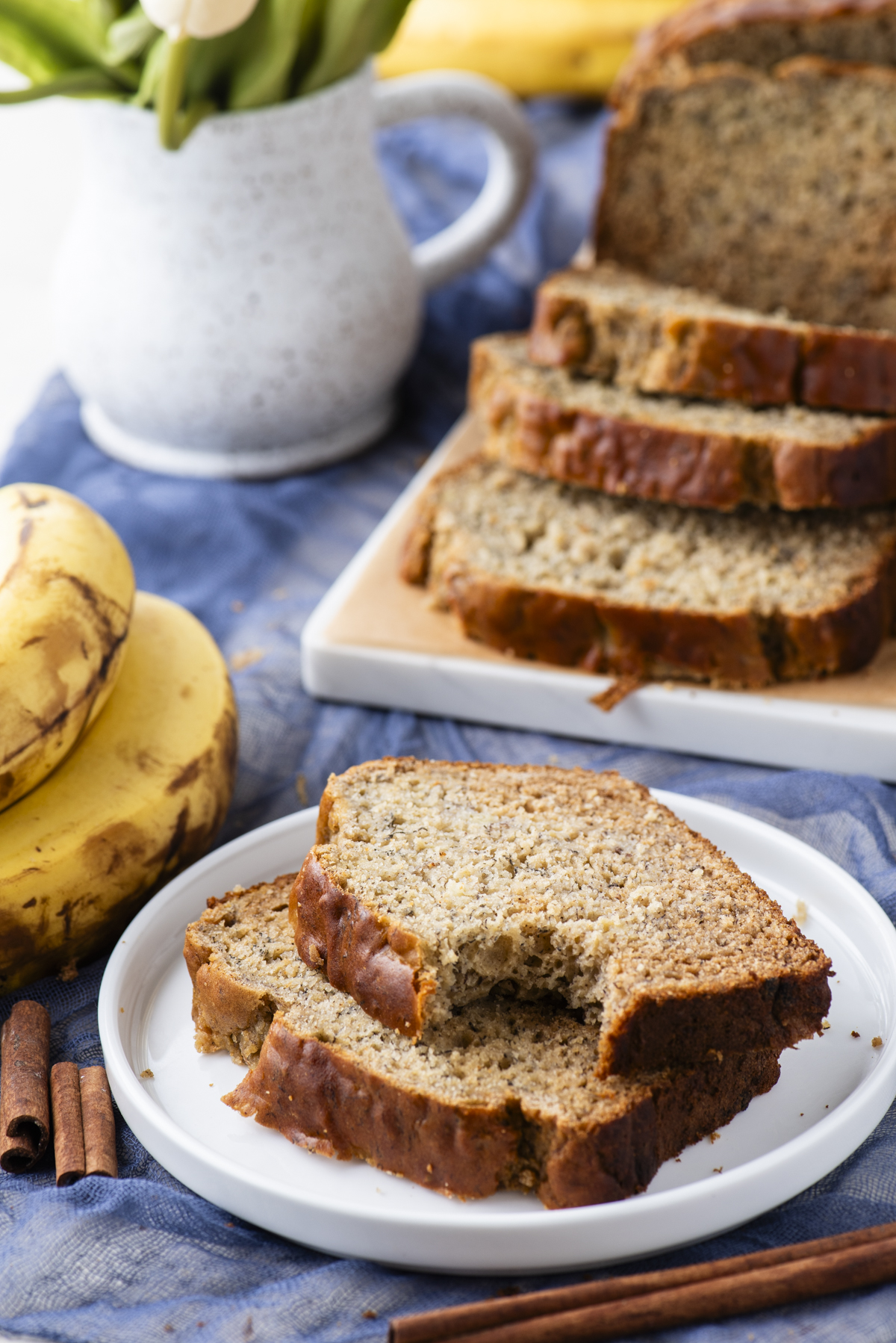 Two slices of banana bread on a white plate in the foreground, with a loaf of sliced banana bread on a cutting board, ripe bananas, cinnamon sticks, and a white pitcher with flowers in the background.