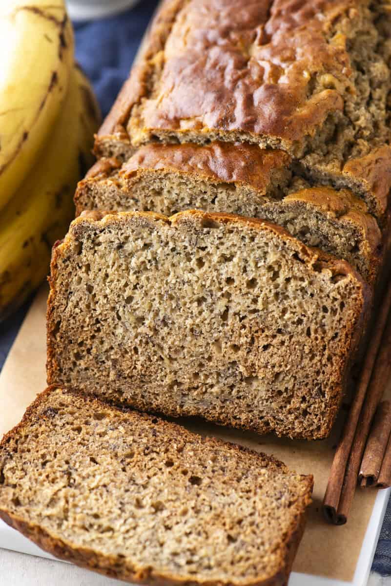 A loaf of banana bread is sliced on a cutting board, with several slices fanned out in front. Ripe bananas and cinnamon sticks are next to the bread. The texture of the bread is moist and slightly dense.
