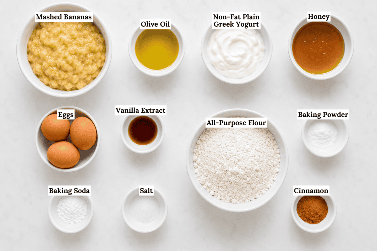 Top-down view of labeled bowls containing mashed bananas, olive oil, non-fat plain Greek yogurt, honey, eggs, vanilla extract, all-purpose flour, baking powder, baking soda, salt, and cinnamon on a white surface.