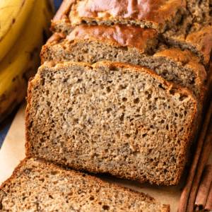 A close-up of sliced banana bread on a wooden board, showing its moist texture. Whole bananas are visible in the background.