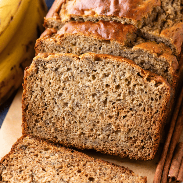 A close-up of sliced banana bread on a wooden board, showing its moist texture. Whole bananas are visible in the background.