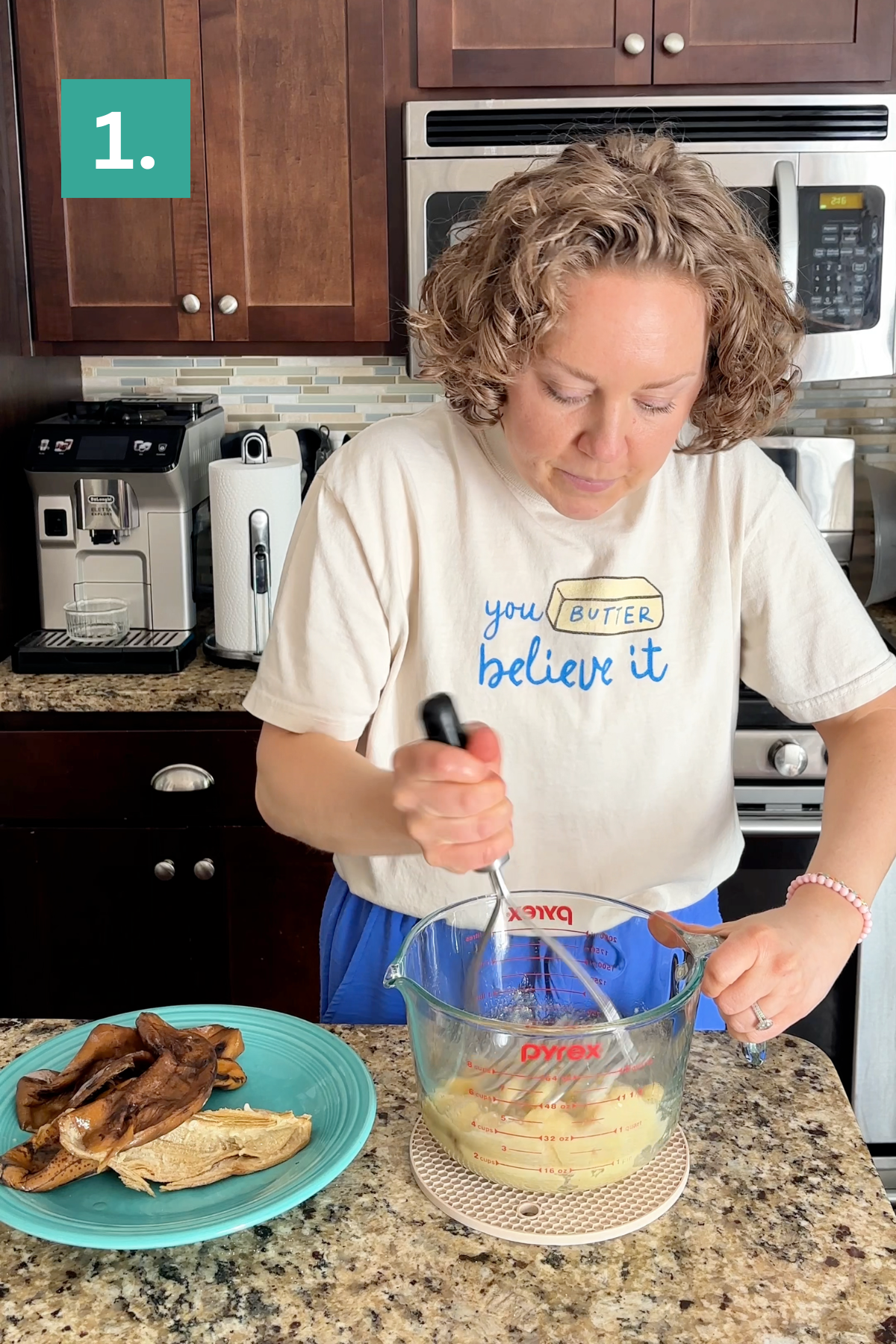 A woman stands in a kitchen mashing potatoes in a glass measuring cup. She wears a shirt that says you BUTTER believe it. Cooked eggplant is on a plate nearby, and coffee machines are in the background.