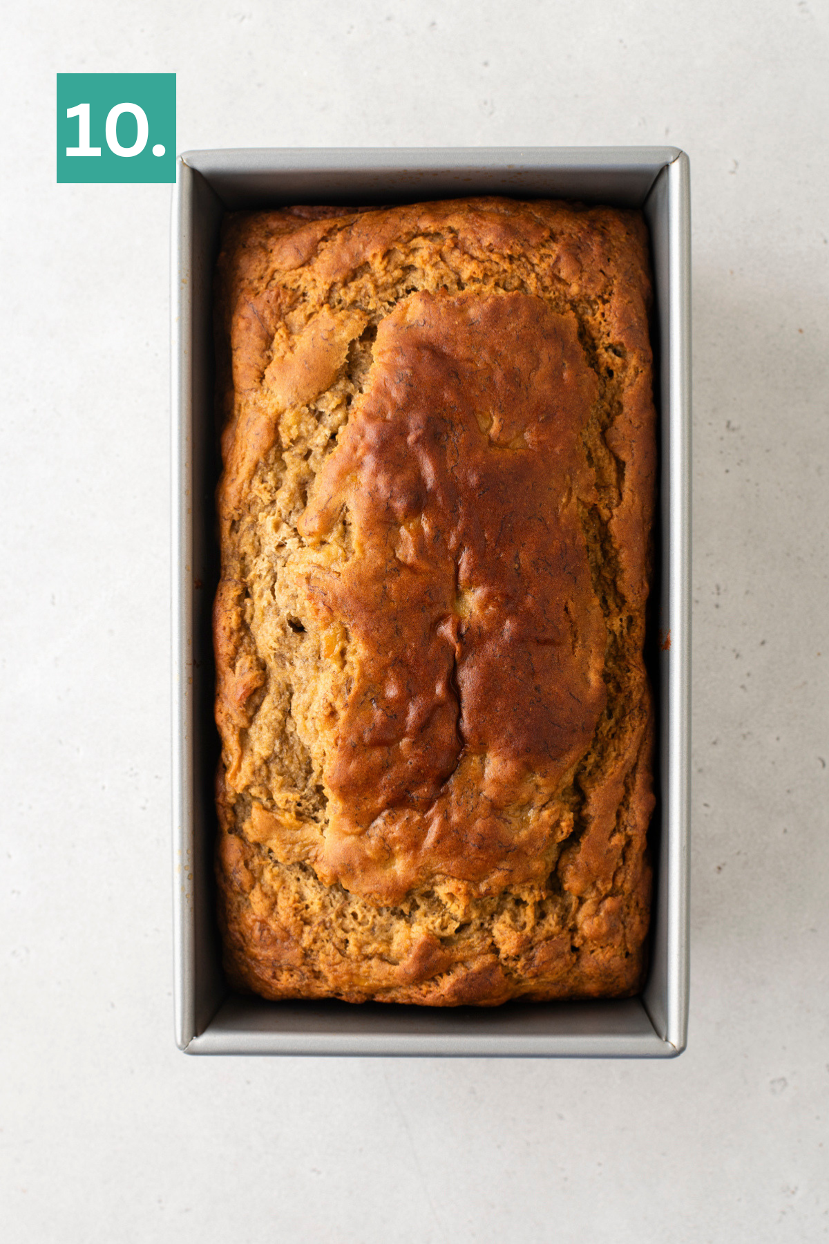 A golden-brown loaf of bread sits in a rectangular metal baking pan on a light surface. A green box with the number 10. is in the top left corner.