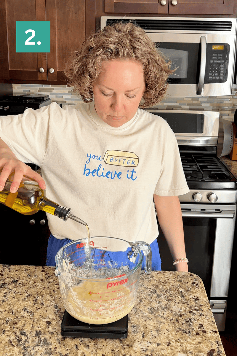 A person pours olive oil into a large Pyrex measuring cup filled with ingredients on a kitchen counter. They wear a shirt that says you BUTTER believe it. A 2. label appears in the top left corner.