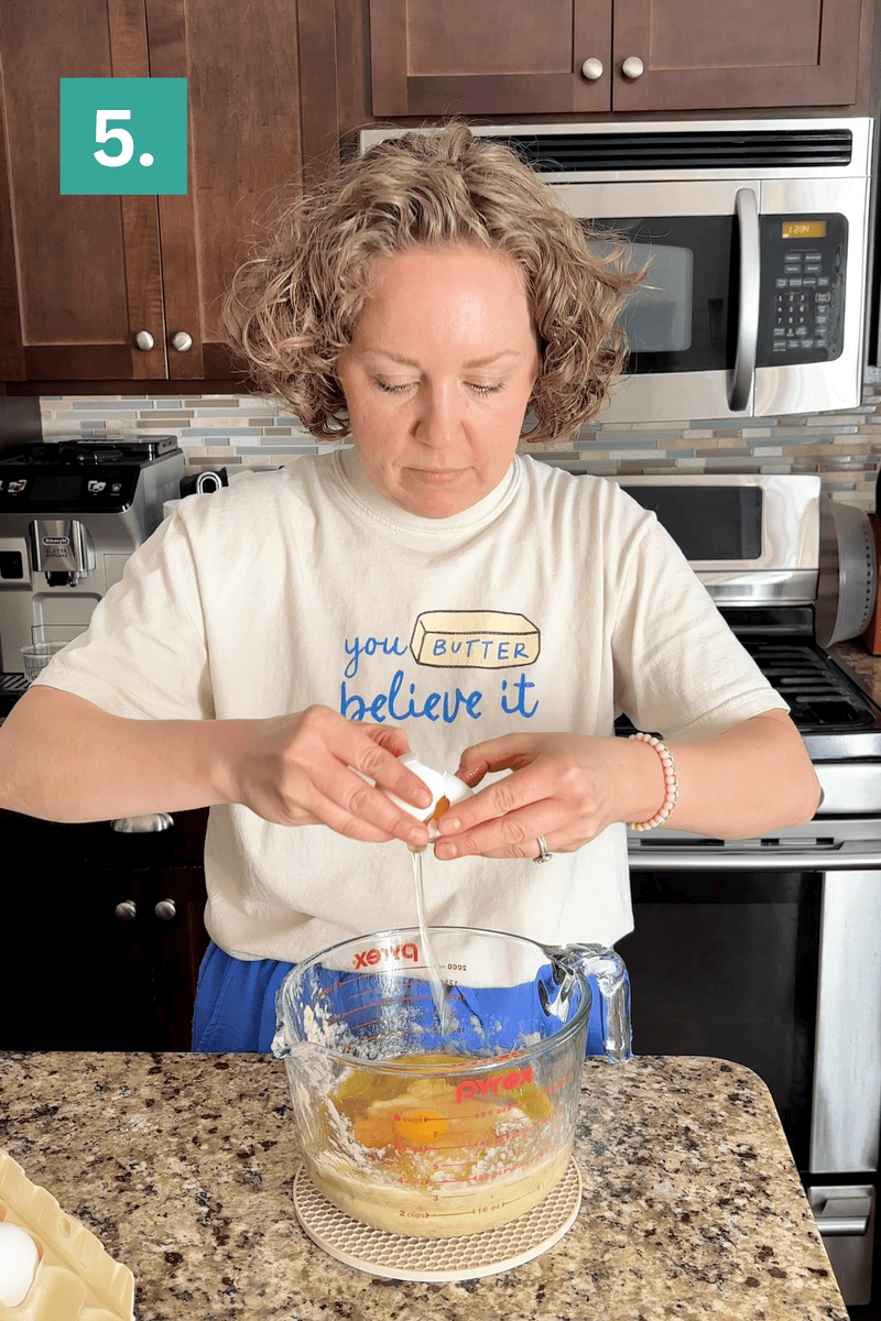 A person cracks an egg into a glass mixing bowl filled with batter in a kitchen. They wear a shirt that says “you BUTTER believe it” and stand in front of a stove and microwave. A green box with “5.” is in the top left corner.
