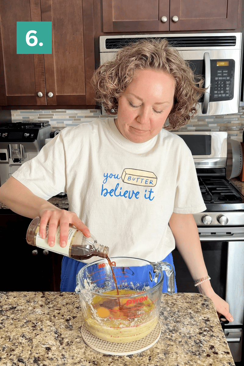 A woman in a kitchen pours vanilla extract from a bottle into a large glass measuring cup with batter. She wears a shirt that says you BUTTER believe it and stands by a stove. A green box with the number 6 is in the top left corner.