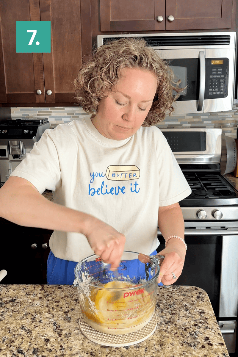 A person with curly hair stirs batter in a glass mixing bowl in a kitchen. She wears a shirt that says “you butter believe it.” A green box with the number 7 is in the top left corner.