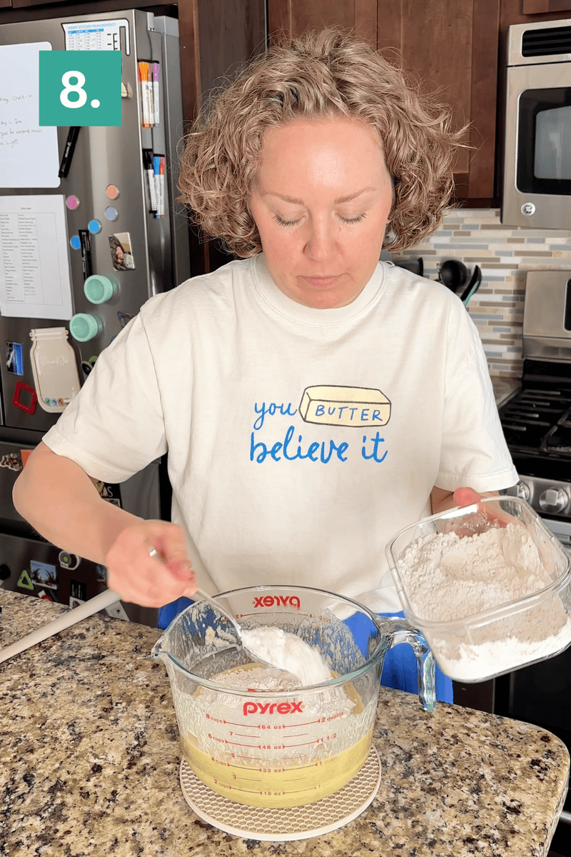 A person in a white “You butter believe it” shirt mixes flour into a Pyrex measuring cup with wet ingredients in a kitchen. A step number “8.” is shown in the top left corner.