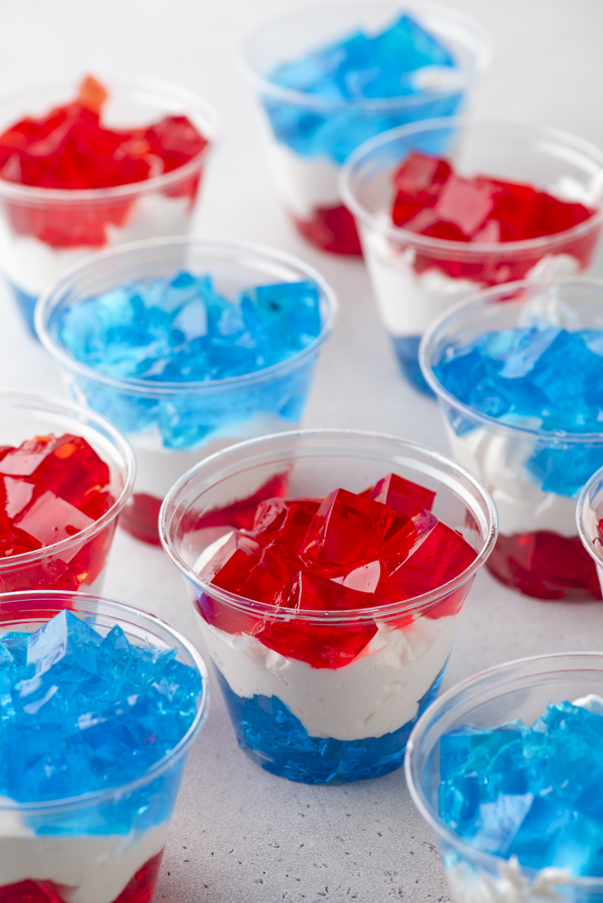 Plastic cups filled with layered red and blue gelatin and white cream, creating a patriotic, tricolor dessert. The cups are arranged on a light surface.