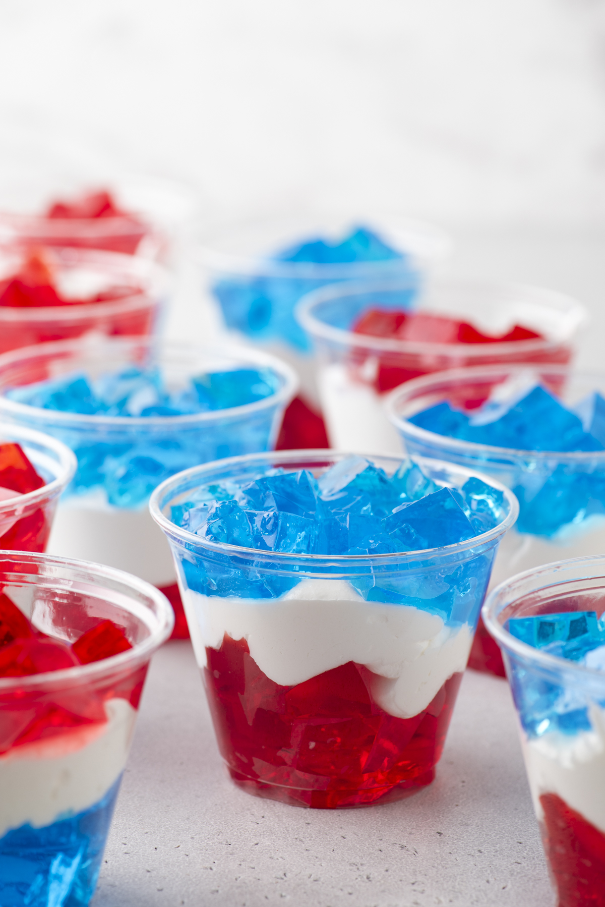 Clear plastic cups filled with layered red and blue gelatin and white creamy filling, arranged on a light surface for a festive, patriotic dessert display.