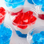 Plastic cups filled with layered blue and red gelatin with a layer of white cream in between, creating a patriotic or festive dessert. Several cups are visible on a light surface.