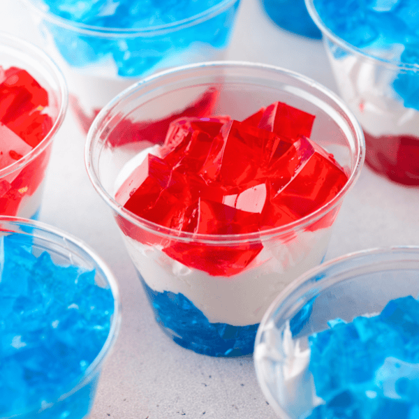 Plastic cups filled with layered blue and red gelatin with a layer of white cream in between, creating a patriotic or festive dessert. Several cups are visible on a light surface.