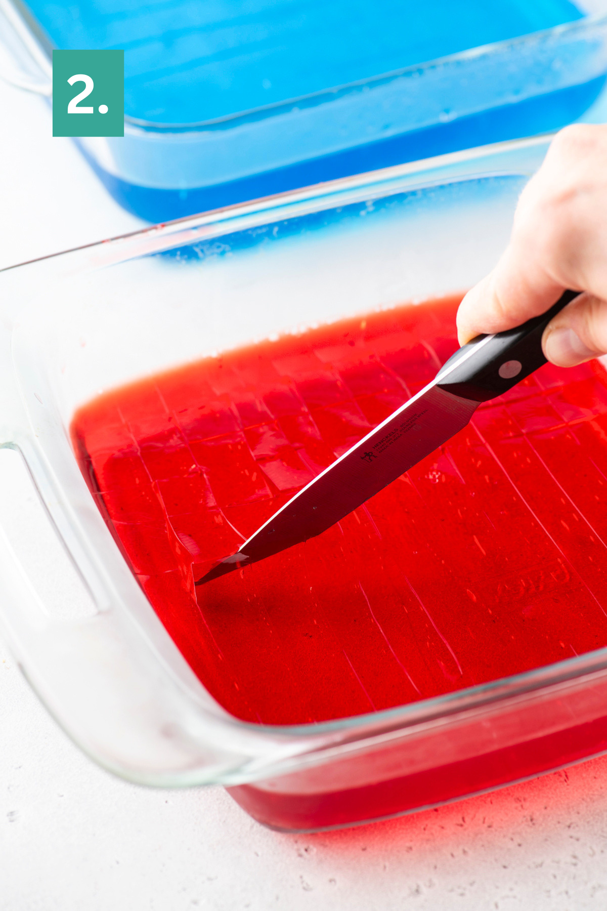 A hand cuts red gelatin in a glass dish with a knife; a dish of blue gelatin sits in the background. A green box with the number 2 is in the top left corner.