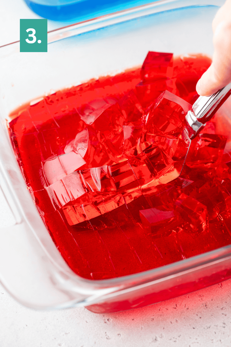 A hand uses a spatula to lift red gelatin cubes from a glass baking dish; a green square with the number 3 is in the upper left corner.