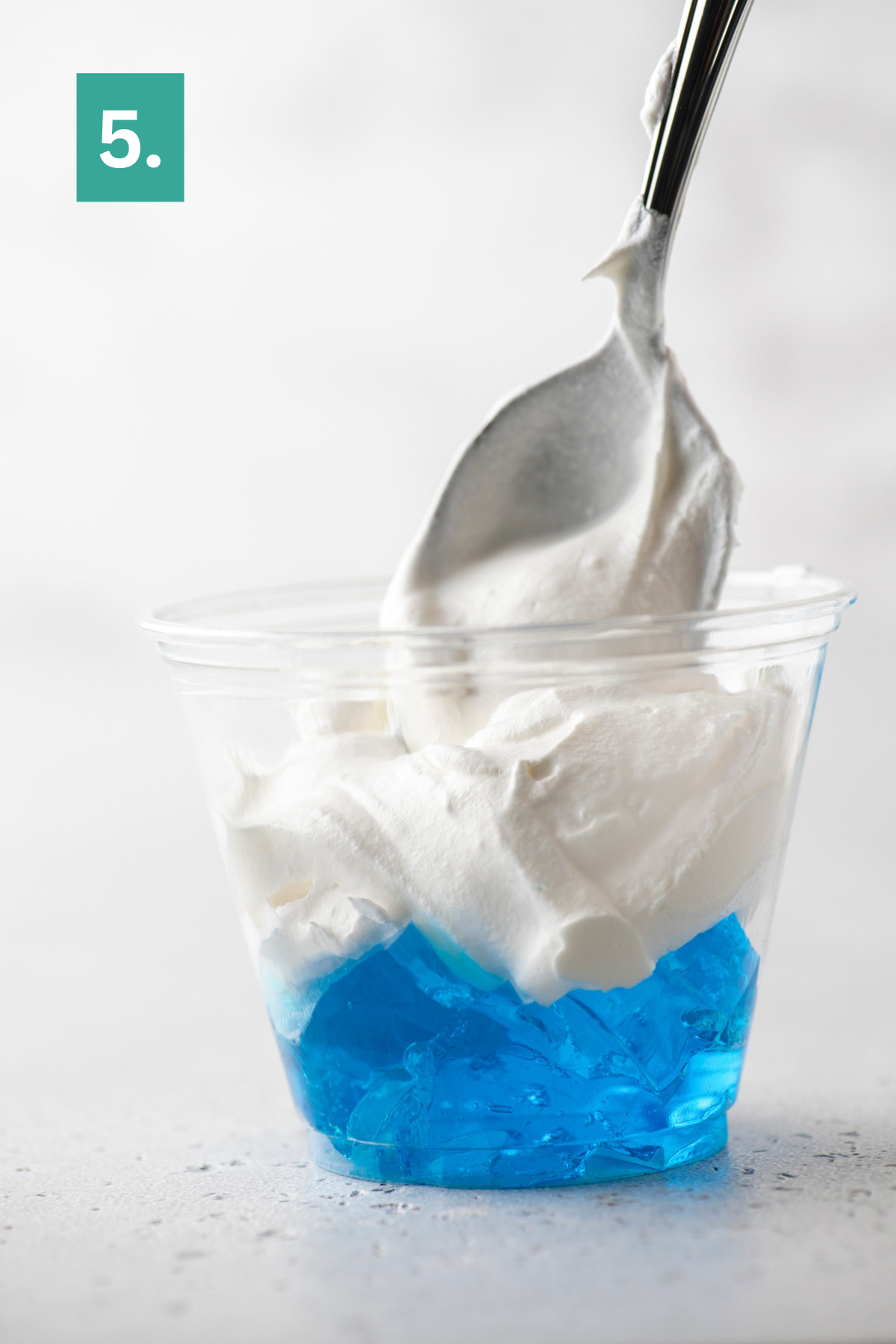 A plastic cup filled with blue gelatin and topped with a generous scoop of whipped cream. A spoon is inserted into the whipped cream. A green box with the number 5 is in the top left corner.