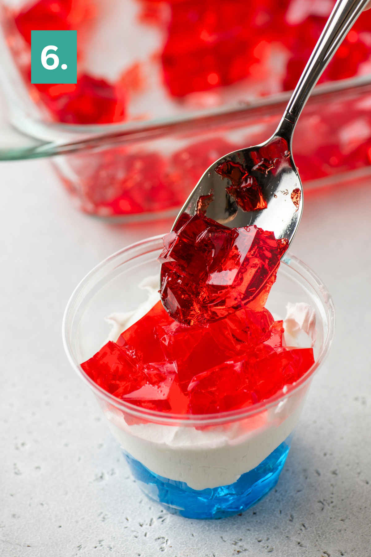 A spoon adds red gelatin cubes on top of whipped cream and blue gelatin in a clear cup. A glass dish with more red gelatin is in the background, and a green box with the number 6 is in the corner.