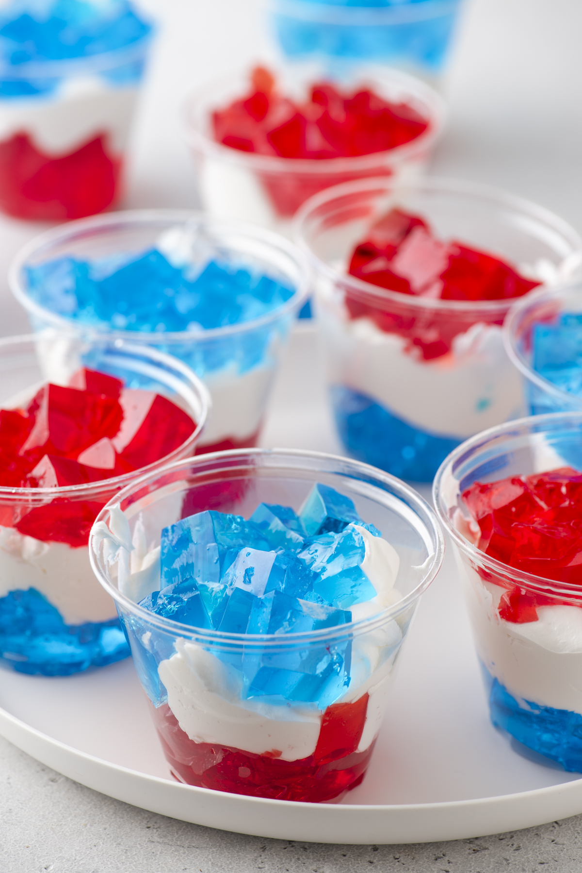 Clear plastic cups filled with layered red and blue gelatin and white whipped cream, arranged on a white tray. The layers create a festive, colorful dessert, suggesting a patriotic or celebratory theme.