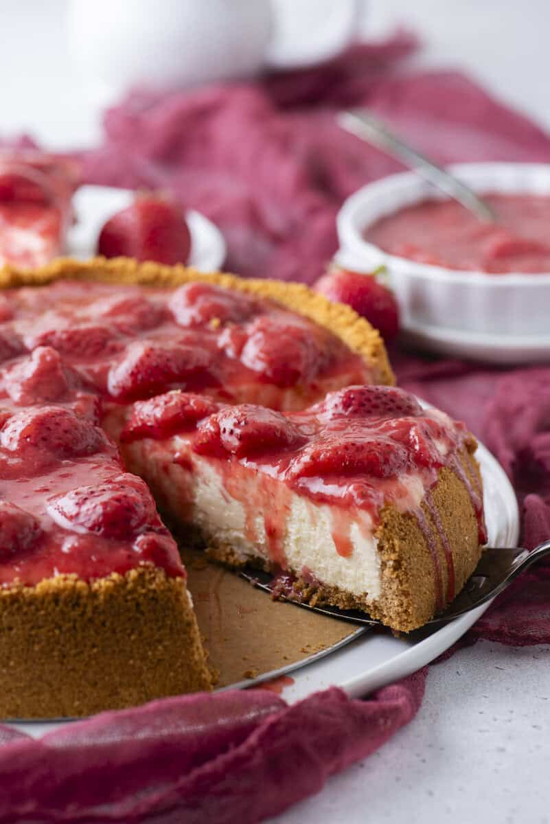 A cheesecake with a golden crust topped with strawberries and strawberry sauce, with one slice removed. A plate with more strawberry sauce and whole strawberries are in the background, all set on a red cloth.