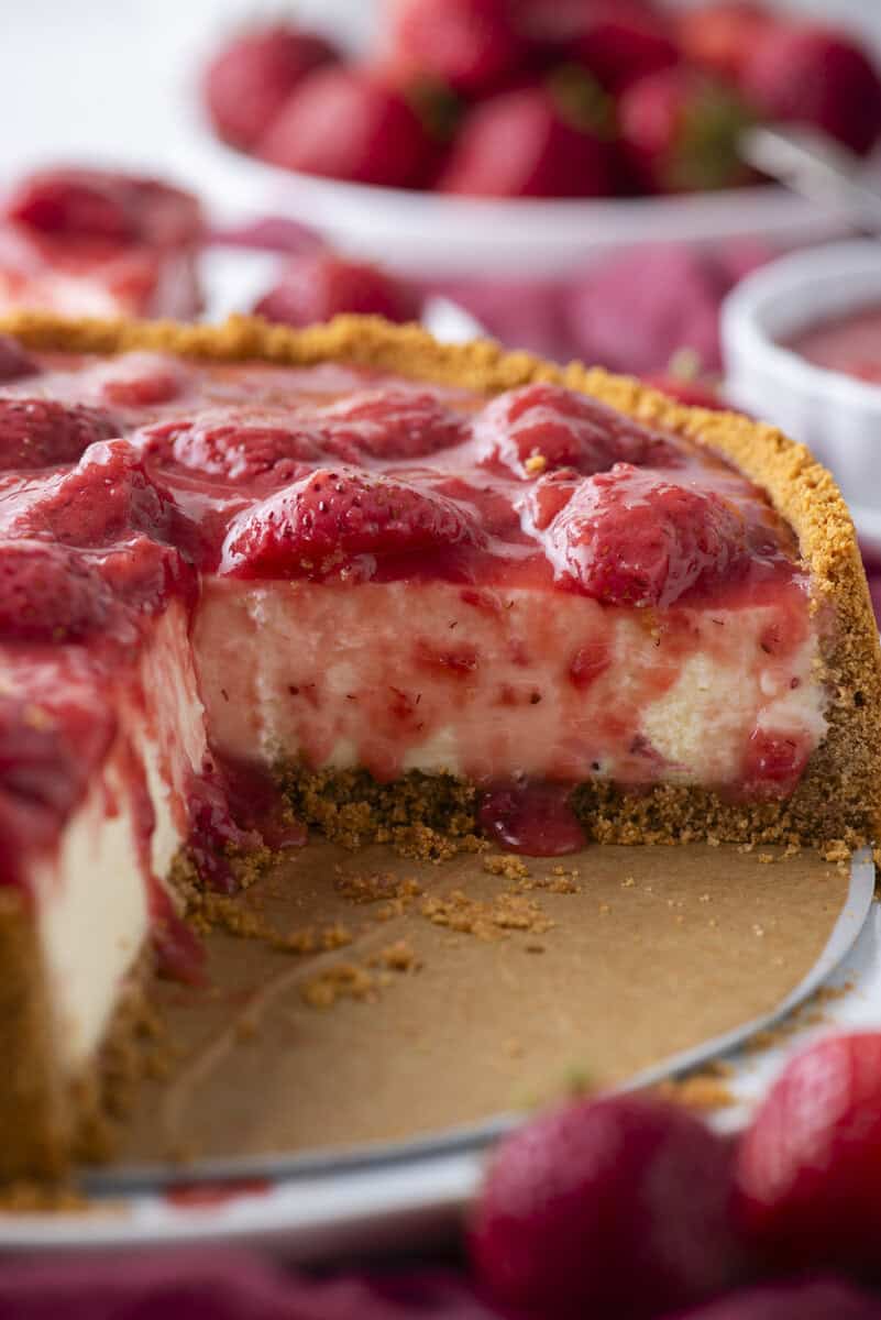 A close-up of a strawberry cheesecake with a graham cracker crust. A slice has been removed, showing creamy filling and strawberry topping. Fresh strawberries are scattered around the cheesecake.