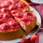 A slice of strawberry cheesecake with a graham cracker crust is being lifted from a plate. The cheesecake is topped with whole strawberries and glossy strawberry sauce. Fresh strawberries are scattered nearby.