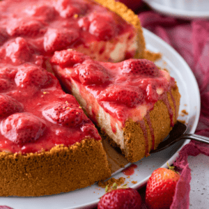 A slice of strawberry cheesecake with a graham cracker crust is being lifted from a plate. The cheesecake is topped with whole strawberries and glossy strawberry sauce. Fresh strawberries are scattered nearby.