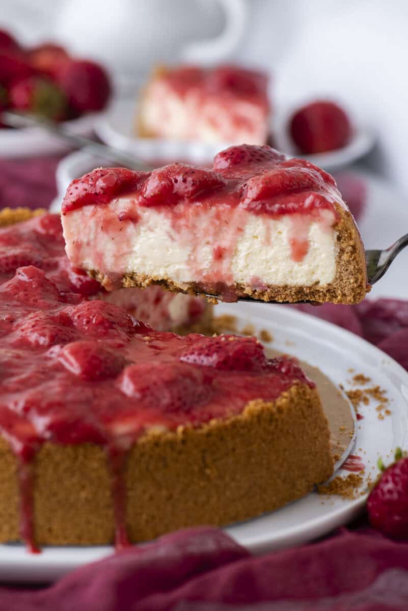 A close-up of a creamy cheesecake topped with strawberry sauce and fresh strawberries. A slice is being lifted from the whole cake, revealing the thick graham cracker crust and smooth filling.